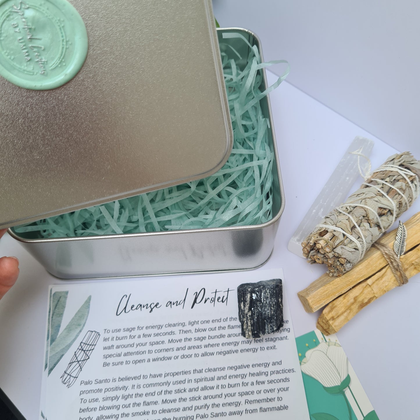 Image of the square tin, information cards, tourmaline, sage and palo santo in background